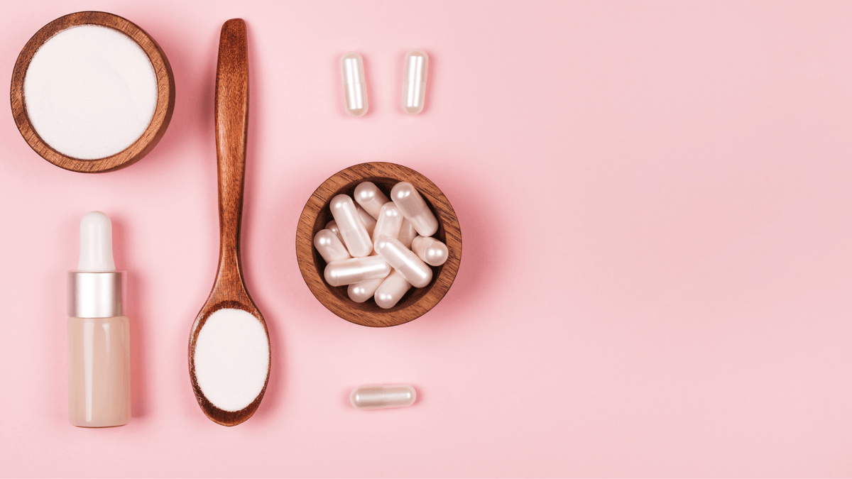 Collagen powder in a wooden bowl and spoon, collagen capsules and a serum bottle on a pink background