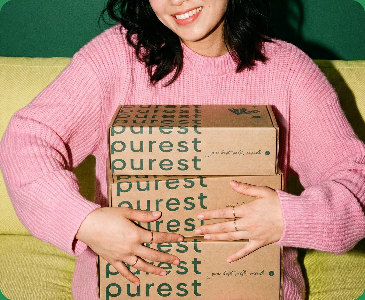 A smiling shopper in a pink jumper hugging a stack of Purest delivery boxes